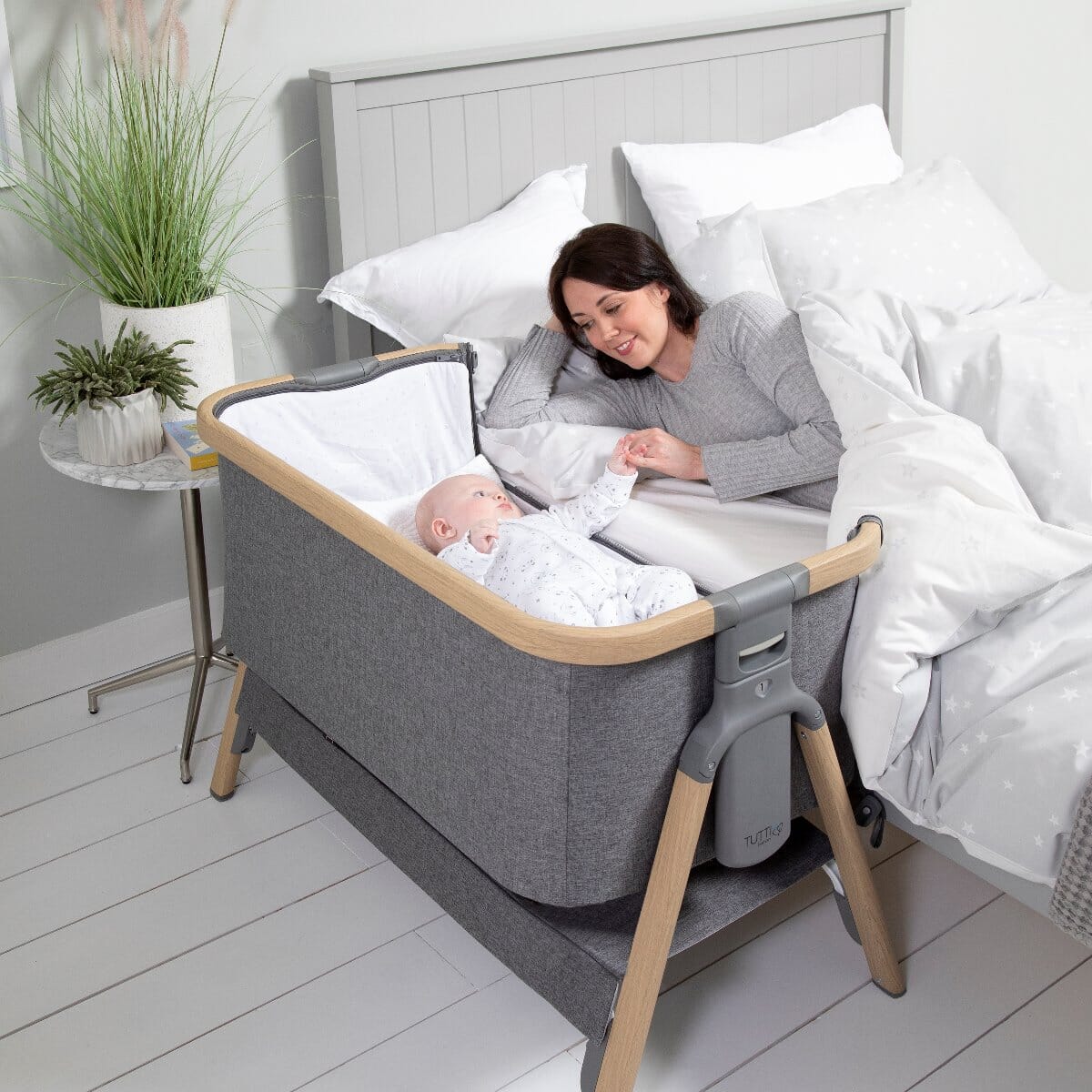 Mum resting beside CoZee Bedside Crib in Oak and Charcoal with baby inside – close, safe co-sleeping setup with mesh side panel visible