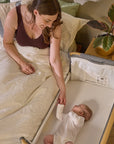 Mother holds baby’s hand while lying beside the CoZee bedside crib in oak and charcoal, highlighting safe co-sleeping design