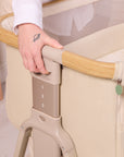 Close-up of the CoZee Air 2 bedside crib in toasted oak/sand showing the adjustable height mechanism with numbered settings.
