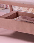 Close-up of CoZee Zen bedside crib in blush showing lower storage shelf with folded muslins and floral bedding.