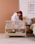 Lifestyle image of a parent settling baby into the CoZee Zen bedside crib in sand, showing ease of access and bedside convenience.