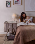 Mother sitting in bed holding baby beside CoZee Sense bedside crib in soft walnut and cocoa finish.