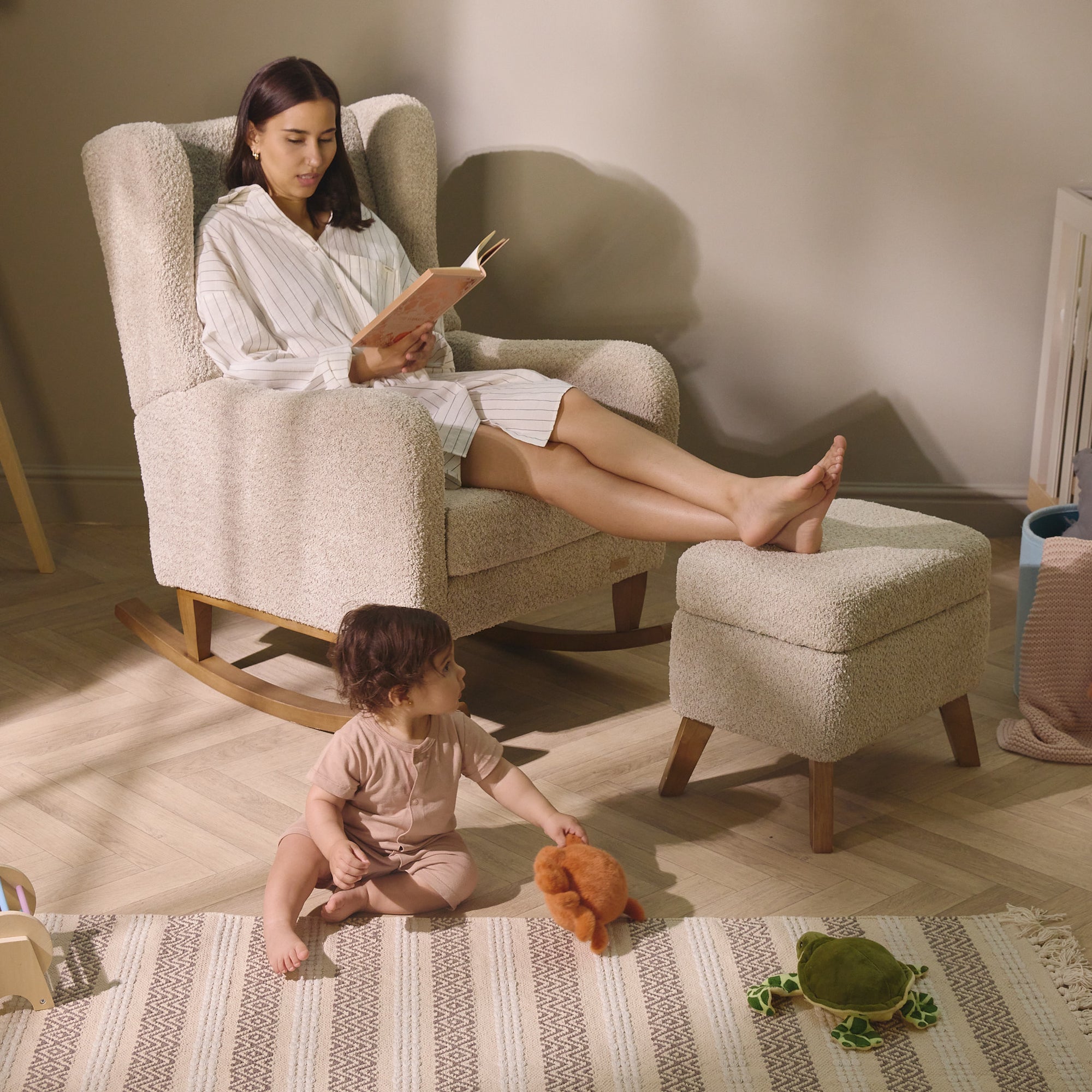 Woman reading in the Harper Wingback rocking chair in teddy mushroom, with footstool and child at play, highlighting comfort and interaction