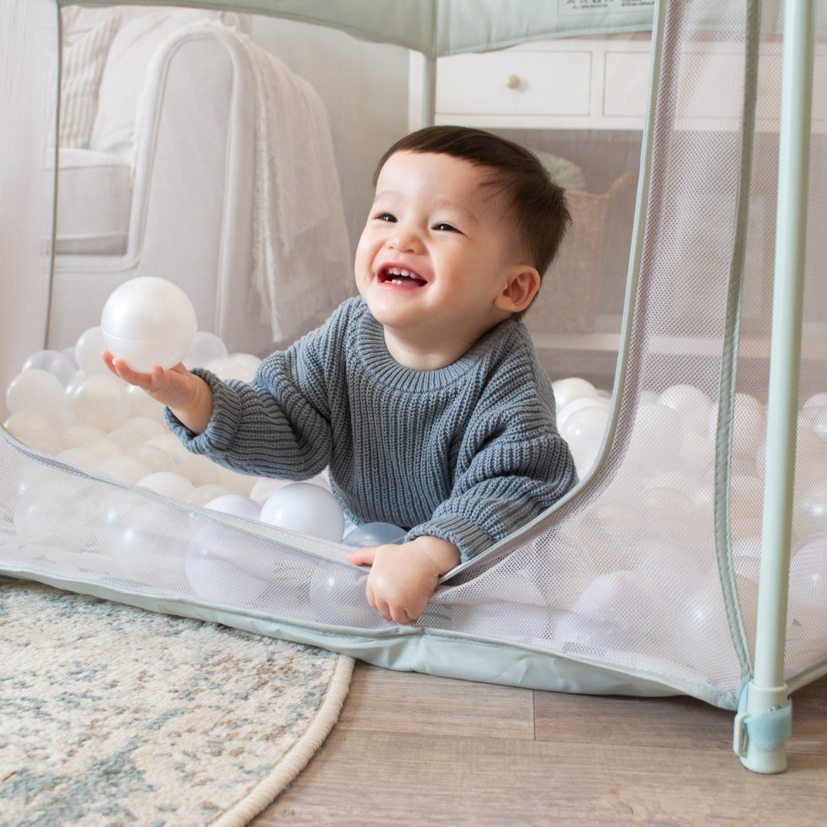 Smiling child leaning on the open side of the Hexa playpen in Leaf Terrazzo, enjoying playtime on the comfy ball-filled base