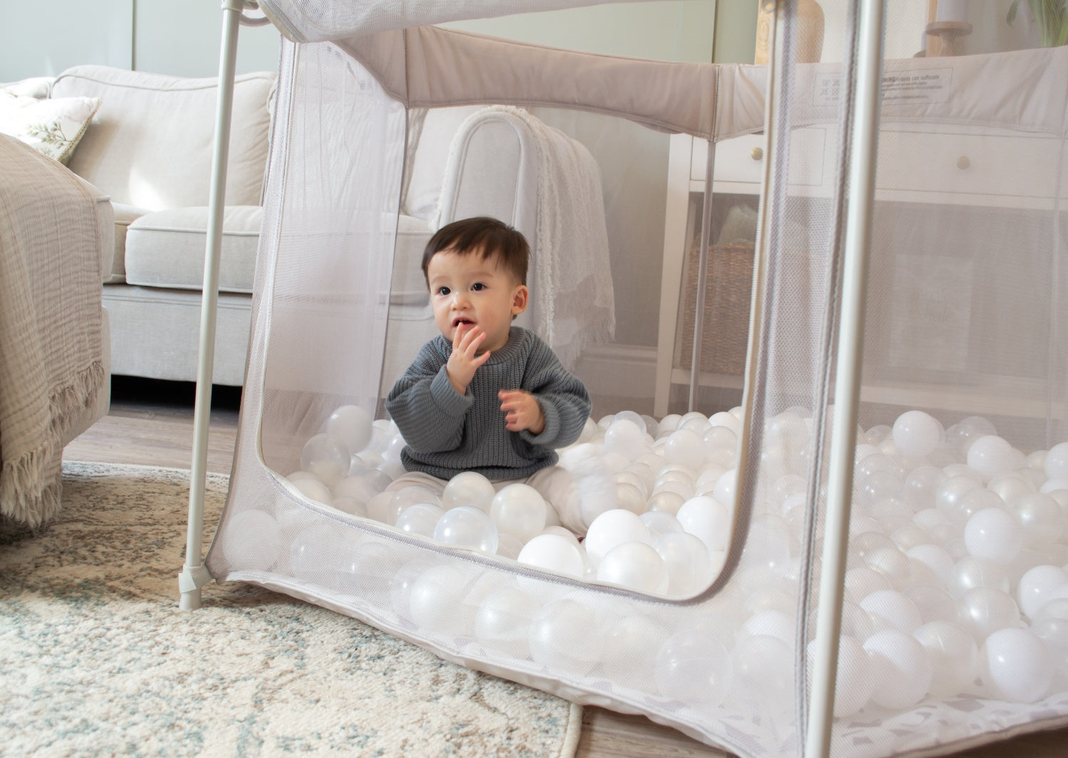Baby sitting inside the Hexa playpen in Mushroom Terrazzo finish, surrounded by white play balls, with zipped mesh side open