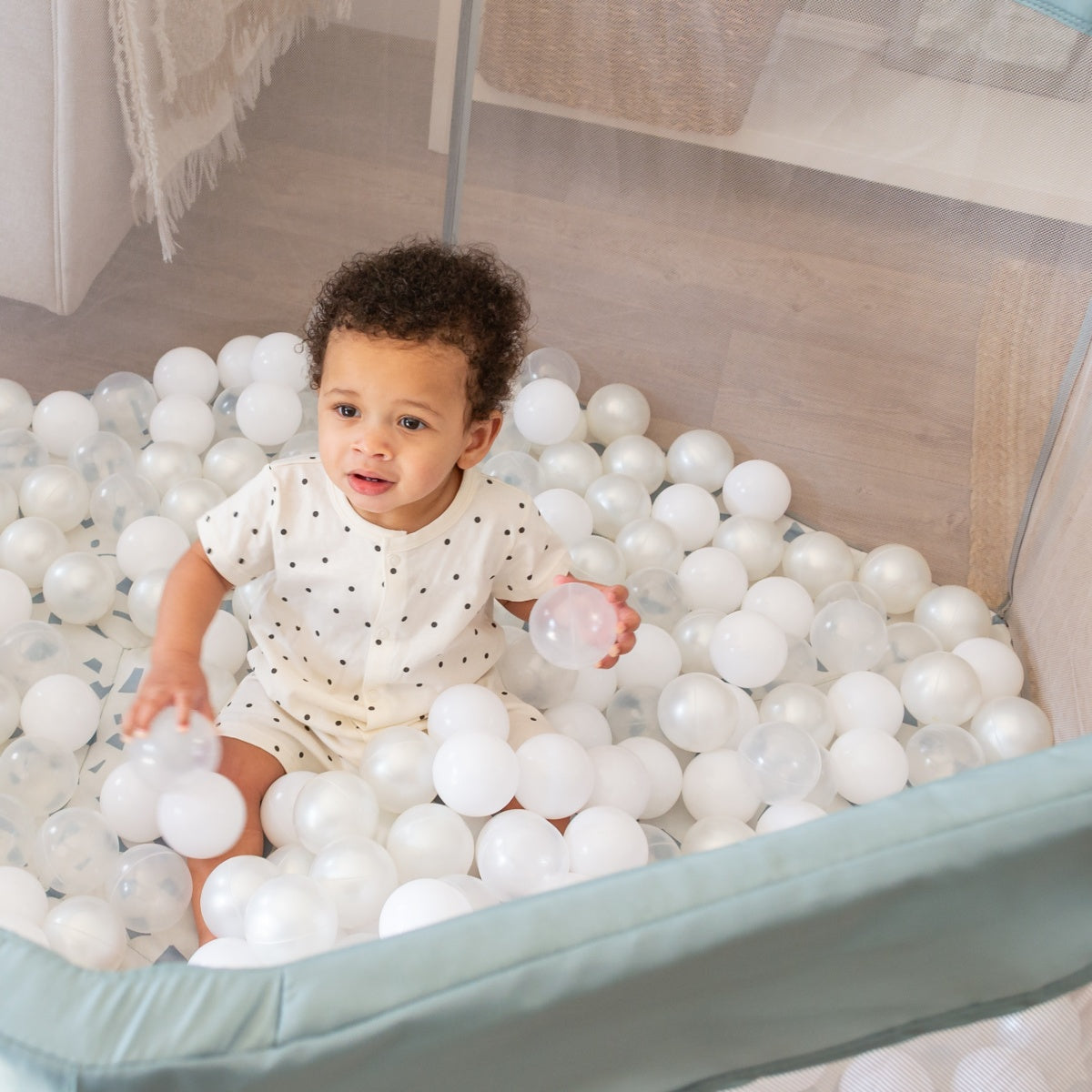 Baby sits inside the Ocean Stone Terrazzo Hexa playpen filled with soft white balls, holding a ball and looking up curiously