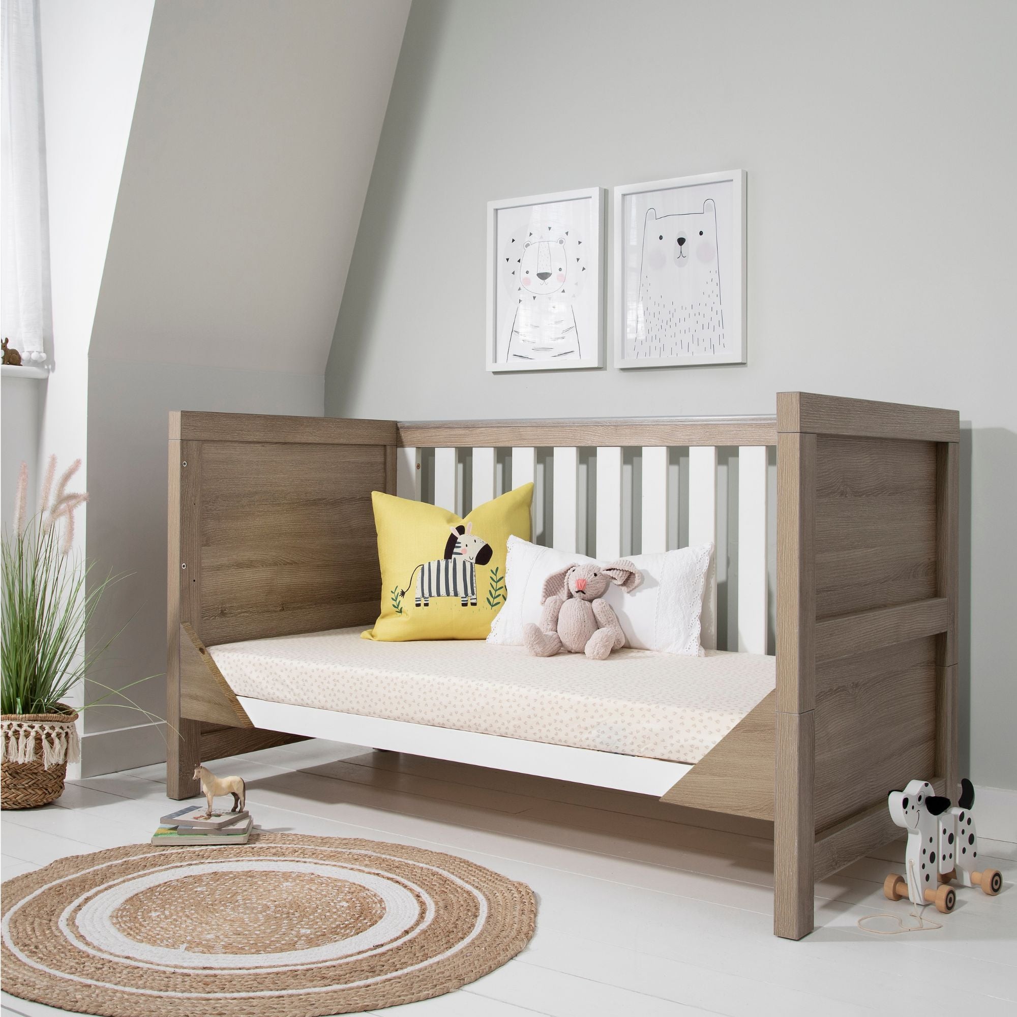Modena Cot Bed in White and Oak shown in sofa bed mode with bedding, cushions, and soft toys, perfect for a modern nursery setting.