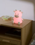 Pink cow-shaped night light on a wooden nightstand with a book next to it.