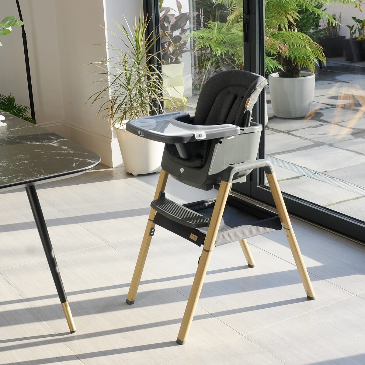 Lifestyle view of Nova Evolutionary Highchair in Grey/Oak placed beside a dining table, showcasing its stylish appearance and functional setup with tray and footrest in a home environment.