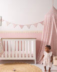 Baby girl standing beside white cot styled with pink bedding, matching tiny tails nursery bunting and soft canopy in a blush nursery.