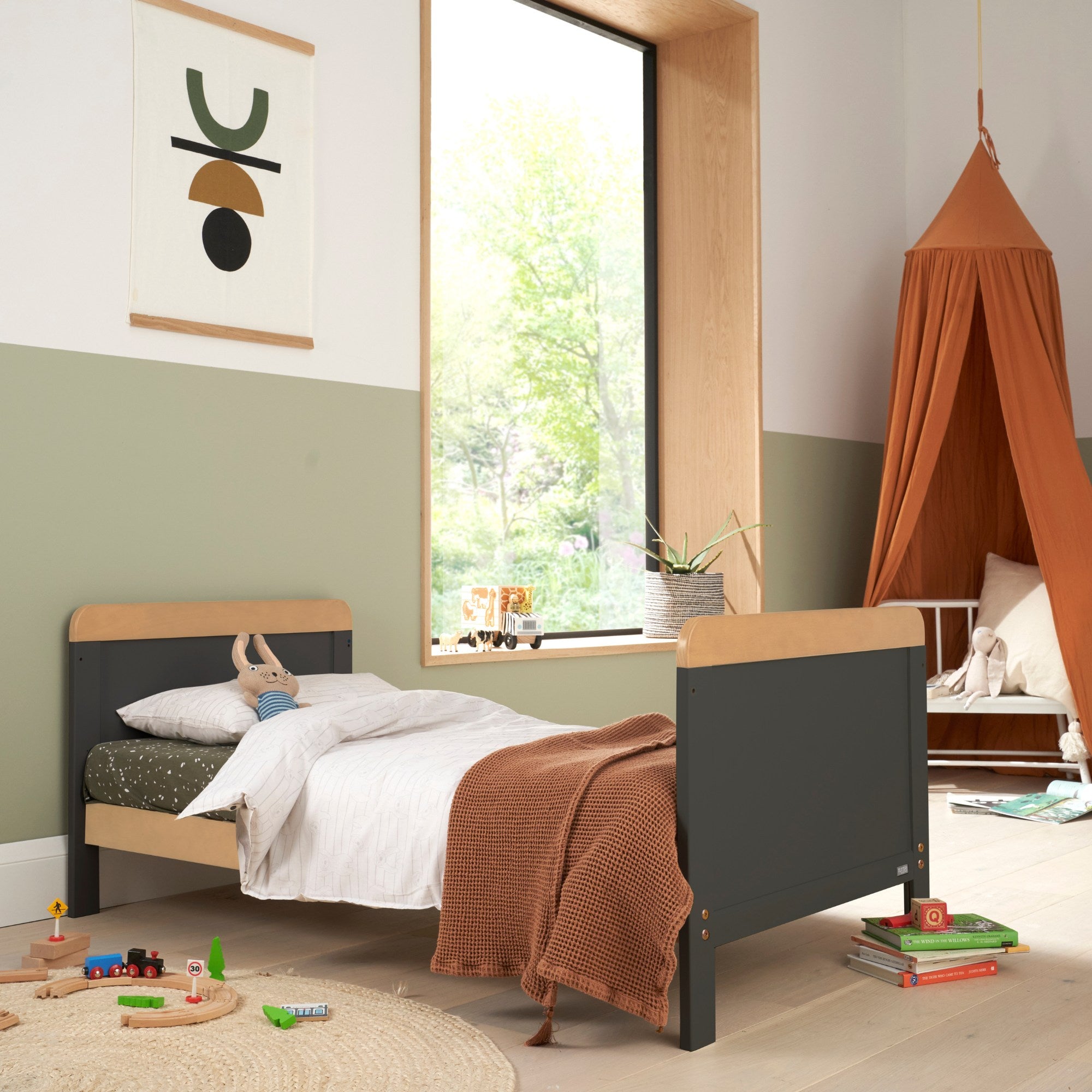 Lifestyle view of the Rio Cot Bed in Slate Grey and Honey Oak in toddler bed mode, styled in a modern nursery with bedding, soft toys, and a canopy.