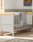 Side view of the Rio cot bed with changing top in Honey Oak and Dove Grey, shown in a nursery setting with a soft green wall, natural light, and plush toys for a cosy and practical environment.