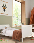 Angled lifestyle view of the Rio cot bed in white converted into a toddler bed styled in a neutral nursery setting.