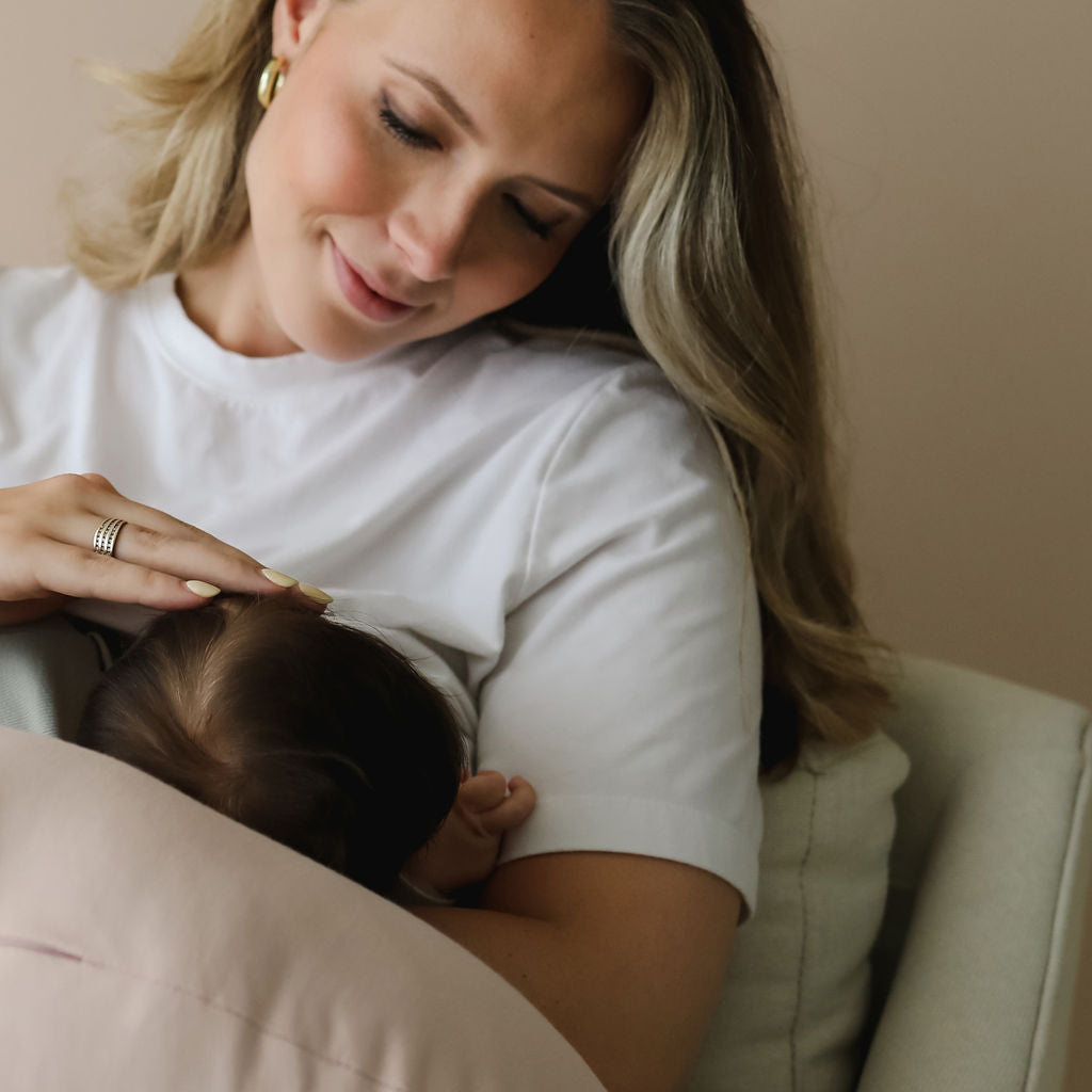 Mother breastfeeding with the SMO nursing pillow in petal, cradling baby comfortably on a armchair