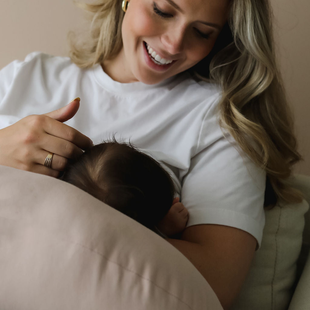 Mother smiling and stroking baby with the SMO nursing pillow in petal