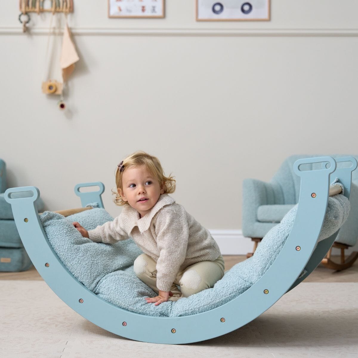 Toddler perched on the Scala Montessori Pikler rocking frame with cushion in boucle ocean stone, exploring and steadying with one hand
