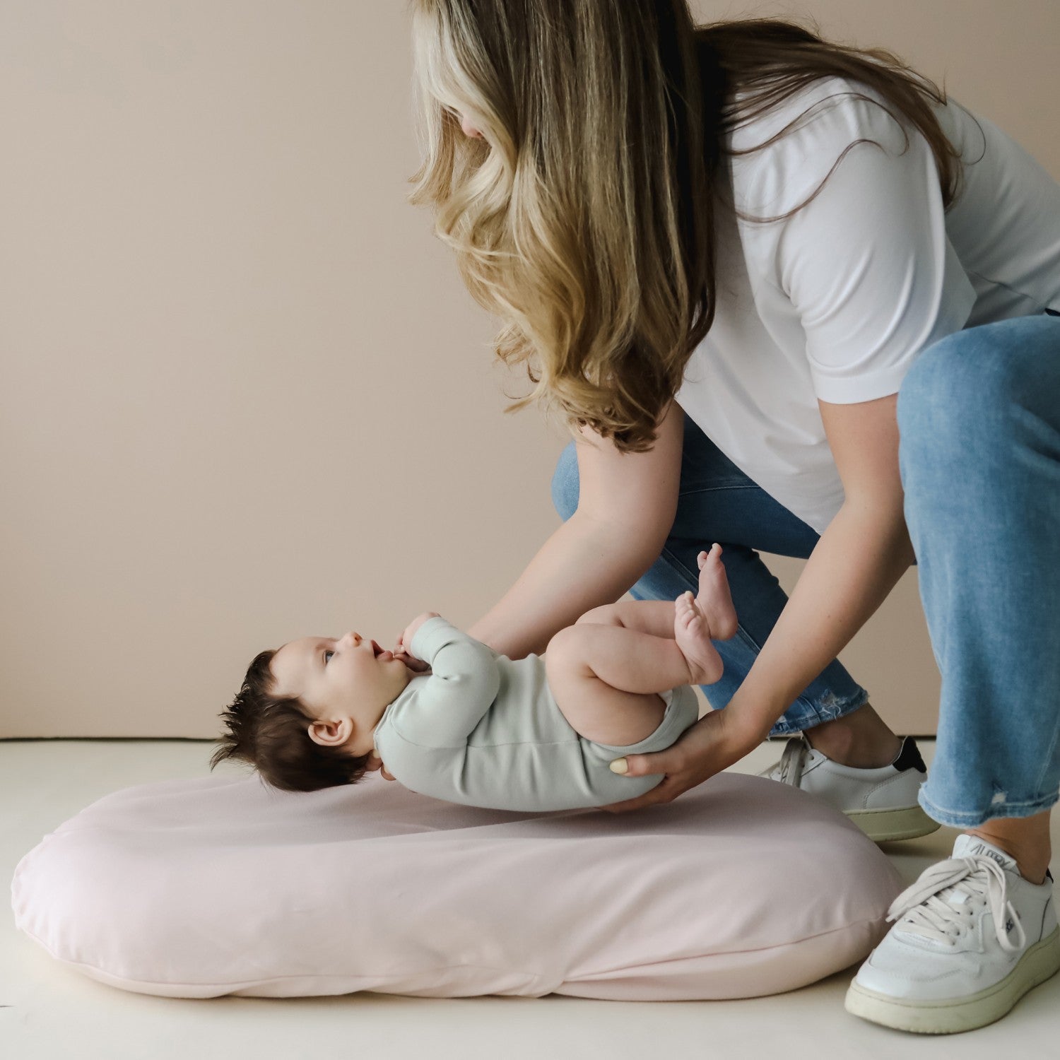A mother gently places her baby into the Snuggle Me lounger with the Petal cover, showcasing the supportive fit and practical daily use
