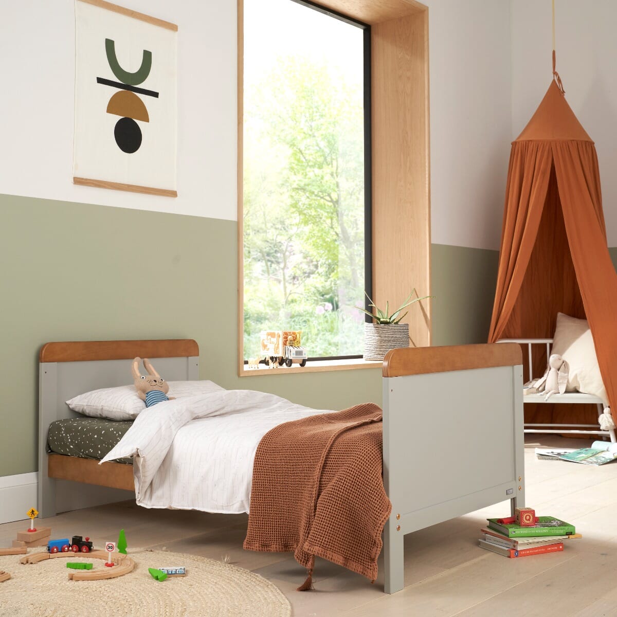 Toddler bed mode of the Rio cot bed in Honey Oak and Dove Grey, styled in a modern nursery with green walls, natural light, playful toys, and a cosy reading nook.