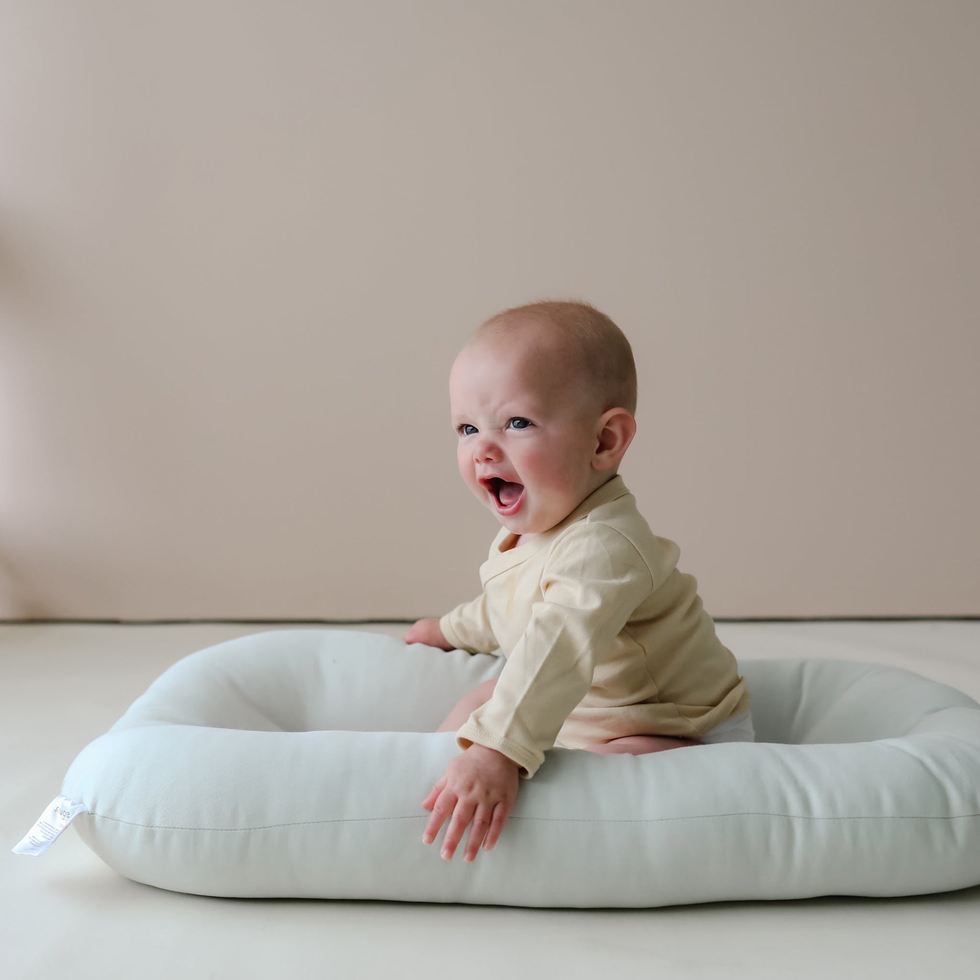 Baby sitting upright in the Snuggle Me Newborn Baby Lounger in Sage, styled in a neutral room with soft lighting and beige backdrop