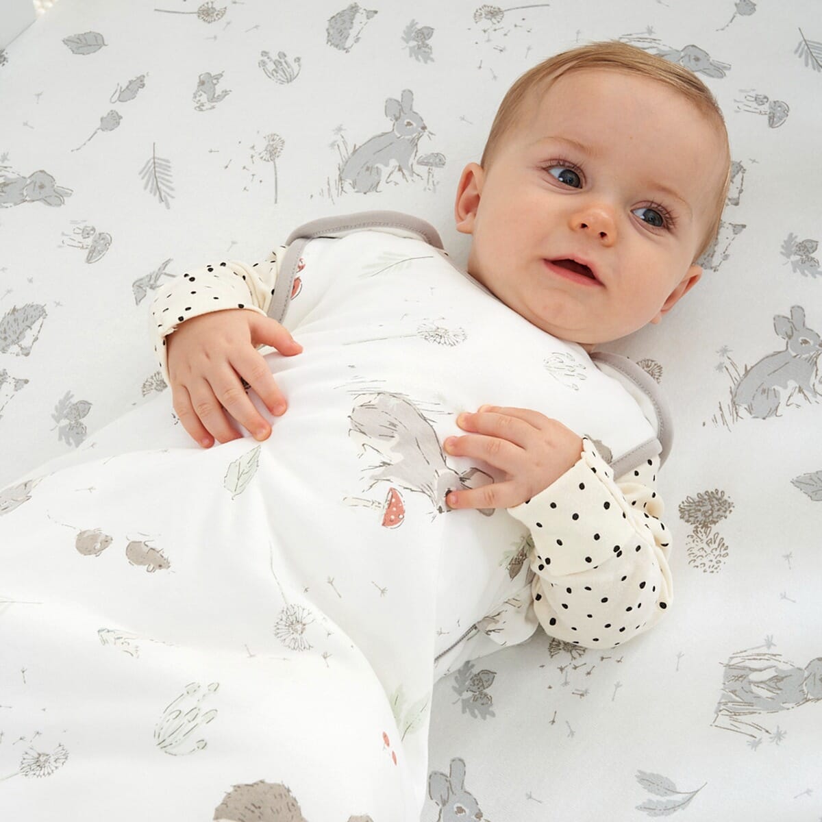 Baby wearing the Cocoon sleep bag 6–18 months, resting on a matching sheet with playful woodland animals in soft neutral tones