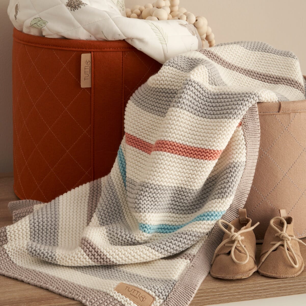 Lifestyle view of Cocoon chunky knitted baby blanket, draped over nursery storage basket, showcasing soft striped cotton knit in cosy tones