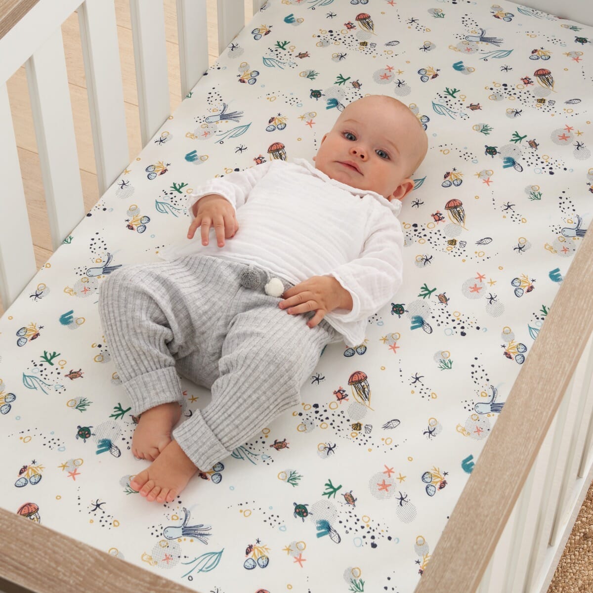 Baby laying on cot bed fitted sheet in Our Planet sea life print, highlighting soft organic cotton fabric for safe, gentle sleep