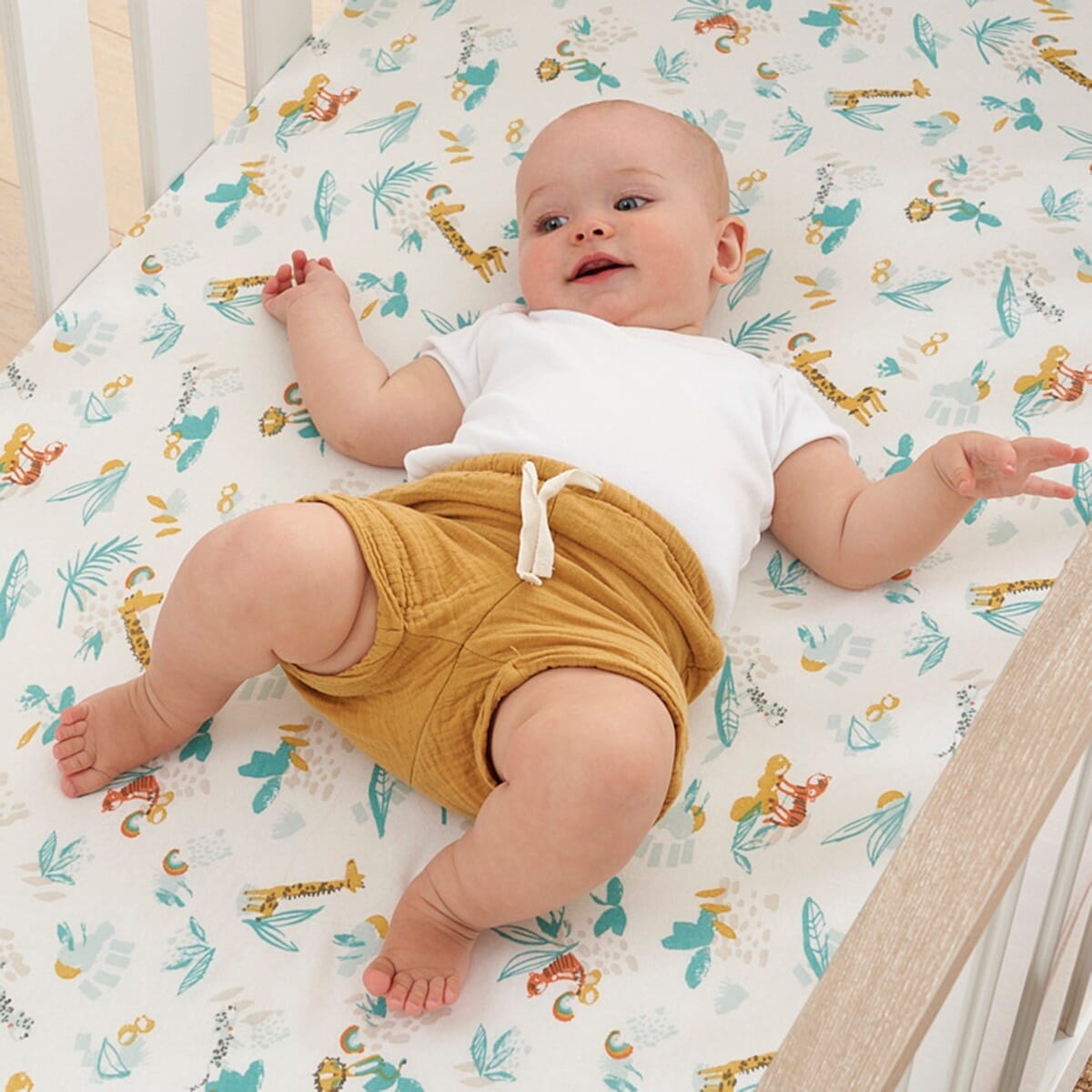 Smiling baby lying on the Run Wild cot bed fitted sheet with safari animal print, adding playful comfort and style to a cosy nursery setting