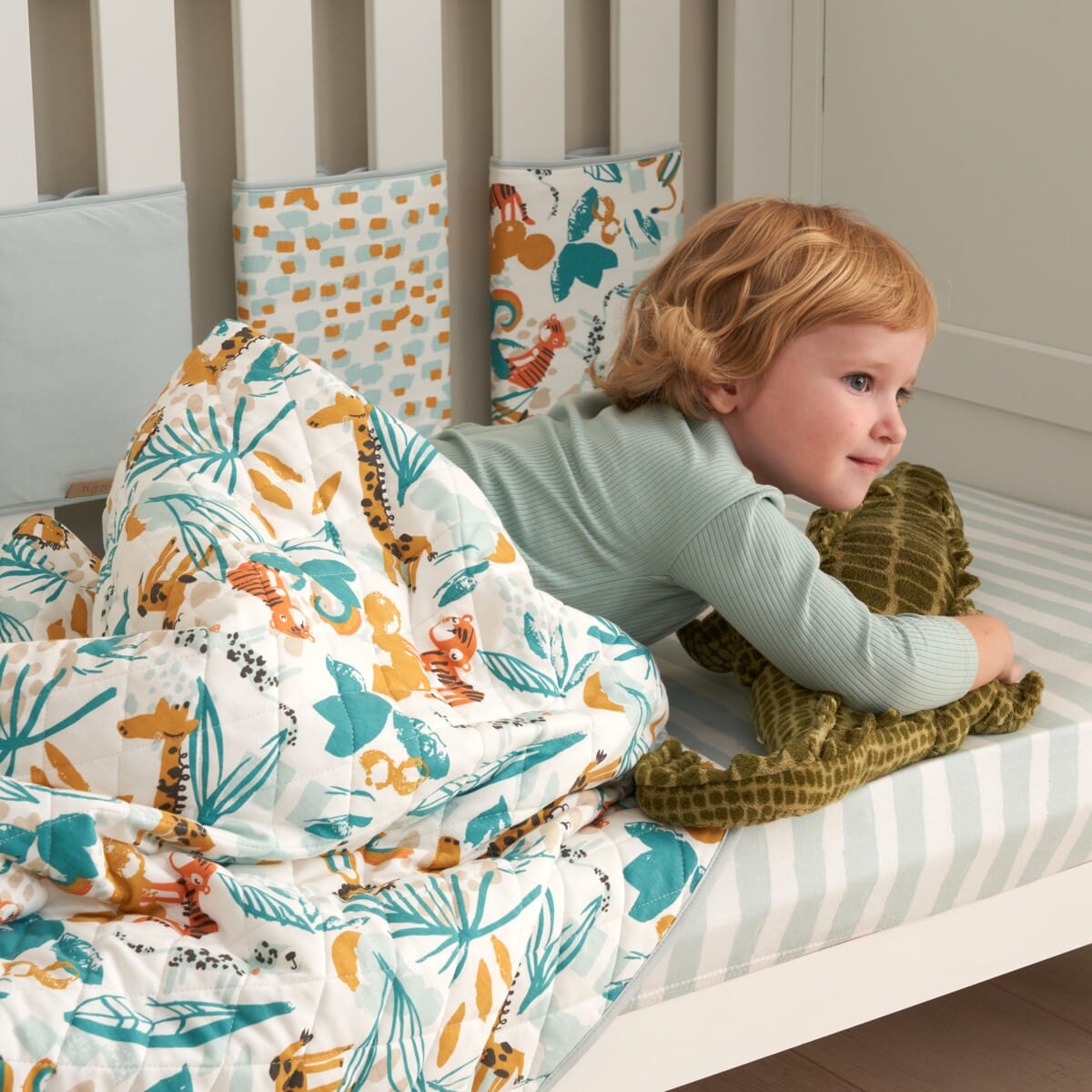 Toddler lying on cot with Run Wild coverlet, quilted cotton with colourful jungle animal prints creating a cosy, playful sleep space
