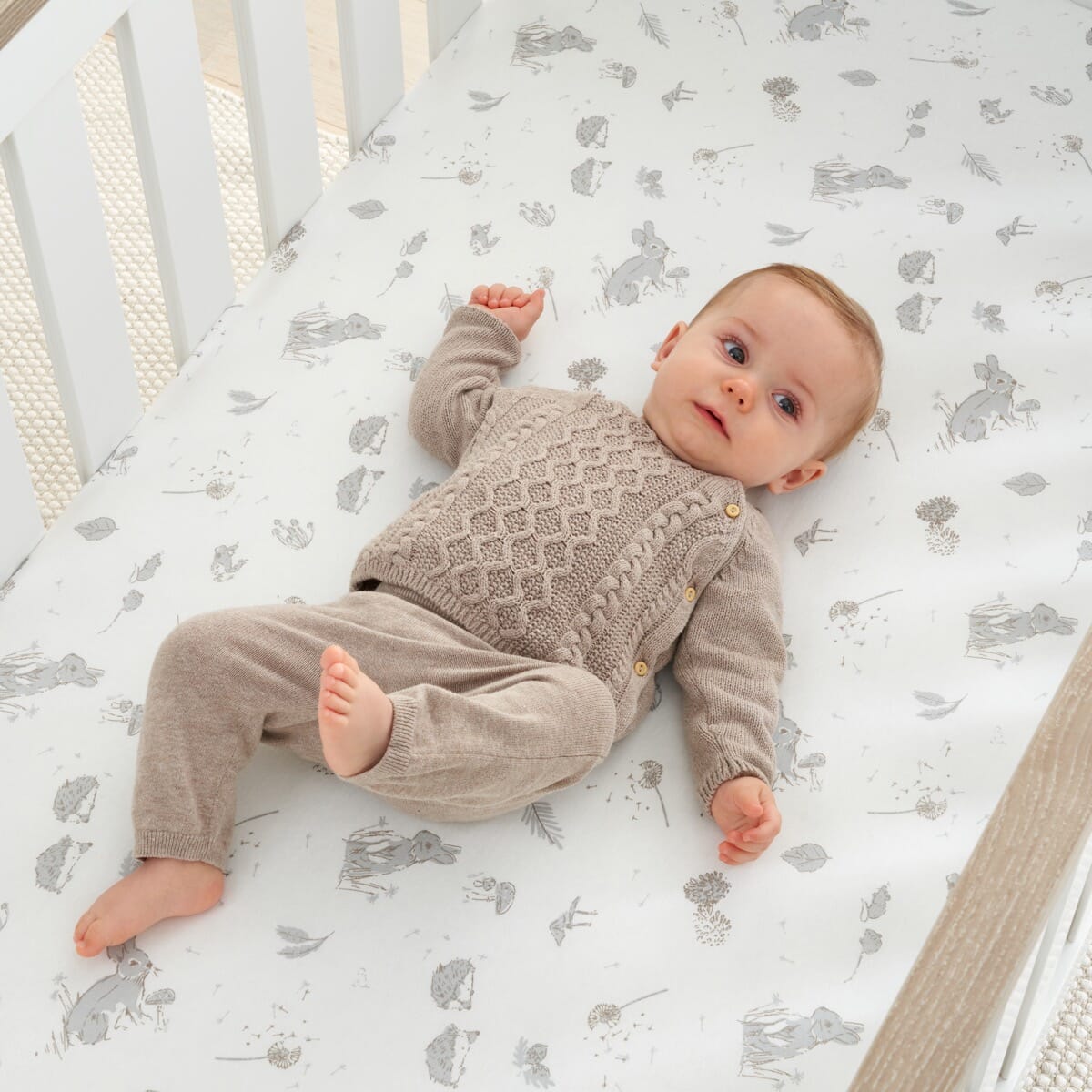 Baby lying on cot fitted sheet in Cocoon design, with woodland animal prints, providing a snug and breathable sleep surface