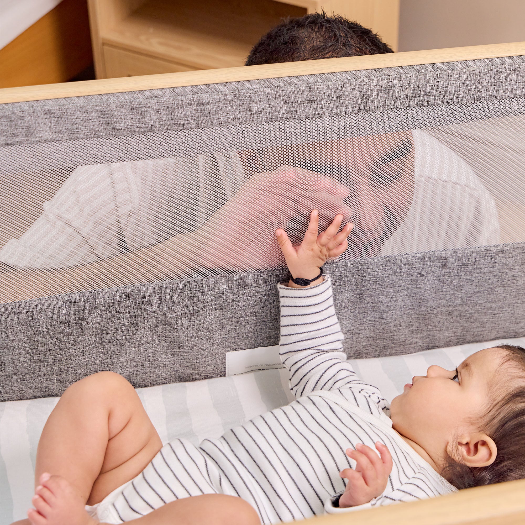Dad looking at baby through mesh side of CoZee Air Bedside Crib in Oak and Charcoal – safe, visible co-sleeping setup