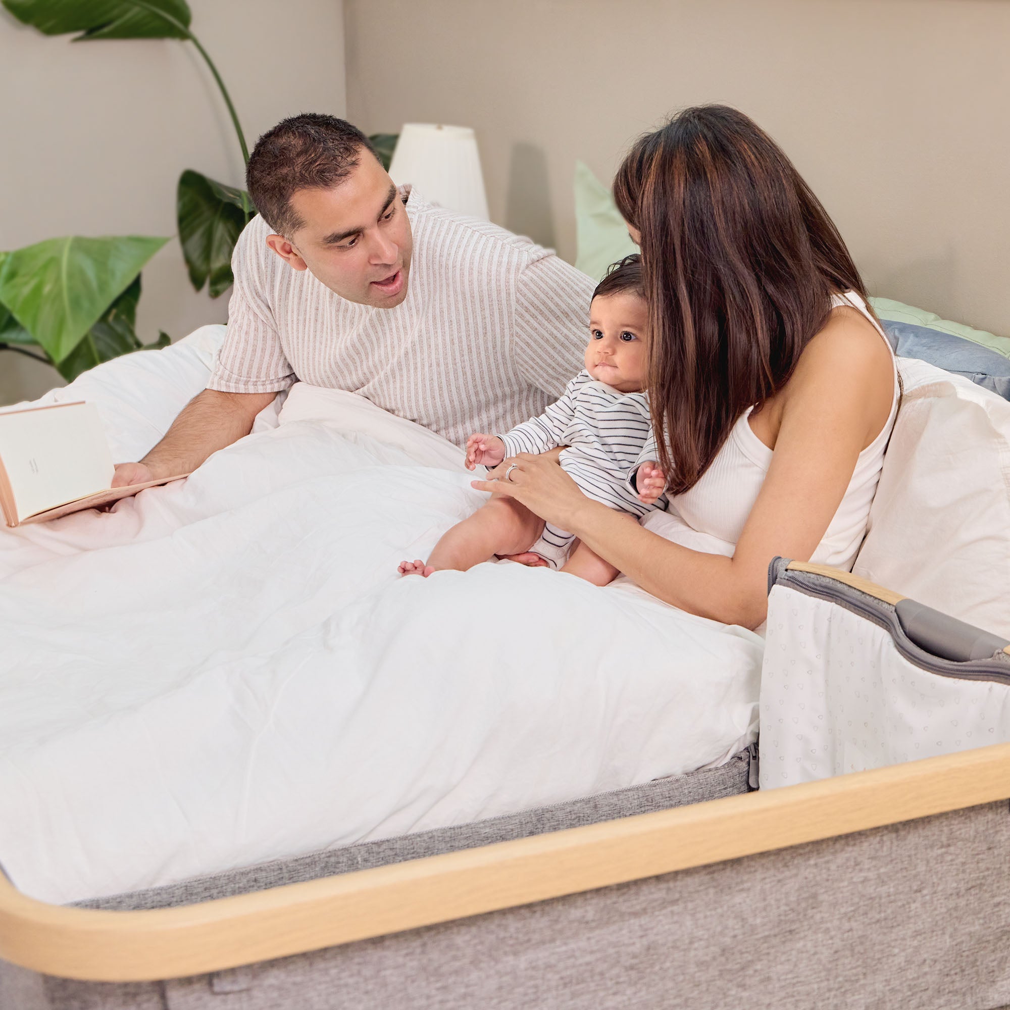 Family relaxing in bed beside the CoZee bedside crib in oak and charcoal, illustrating cosy side-sleeping setup for bonding and reassurance