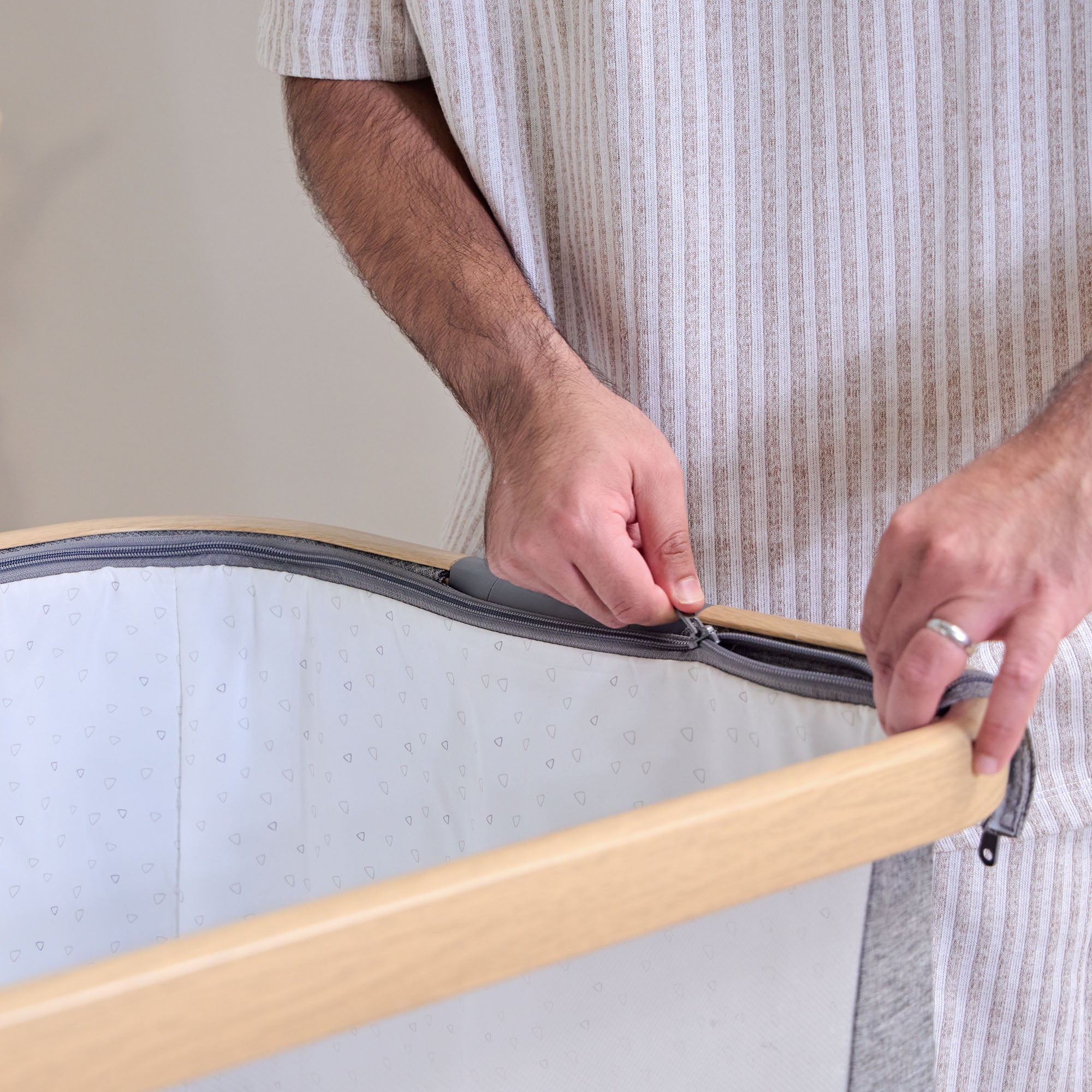 Close-up of a parent zipping the inner liner of the CoZee bedside crib in oak and charcoal, showing easy-access, removable fabric design