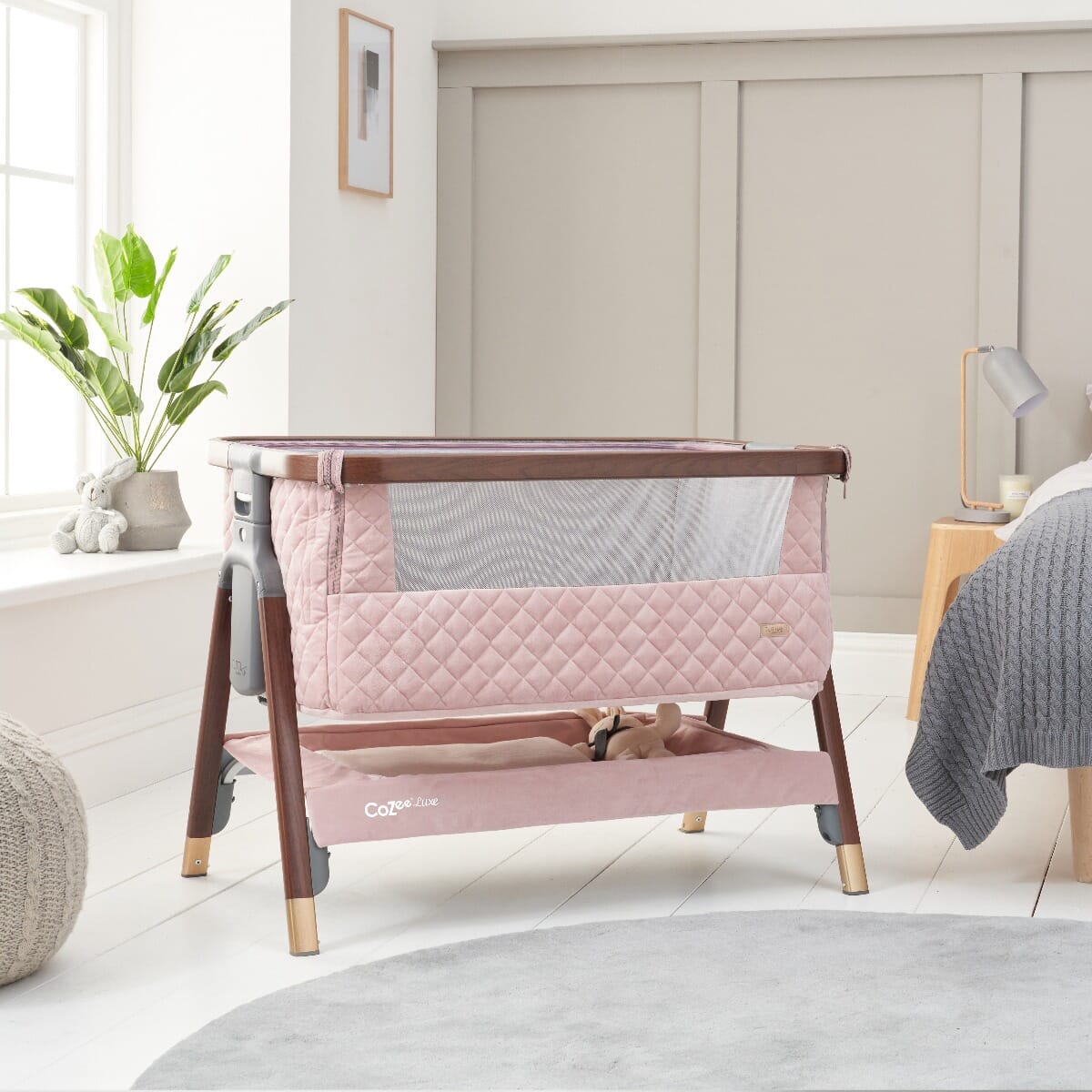 Styled nursery scene featuring the CoZee Luxe bedside crib in walnut and blush with storage shelf and soft mesh side panels.