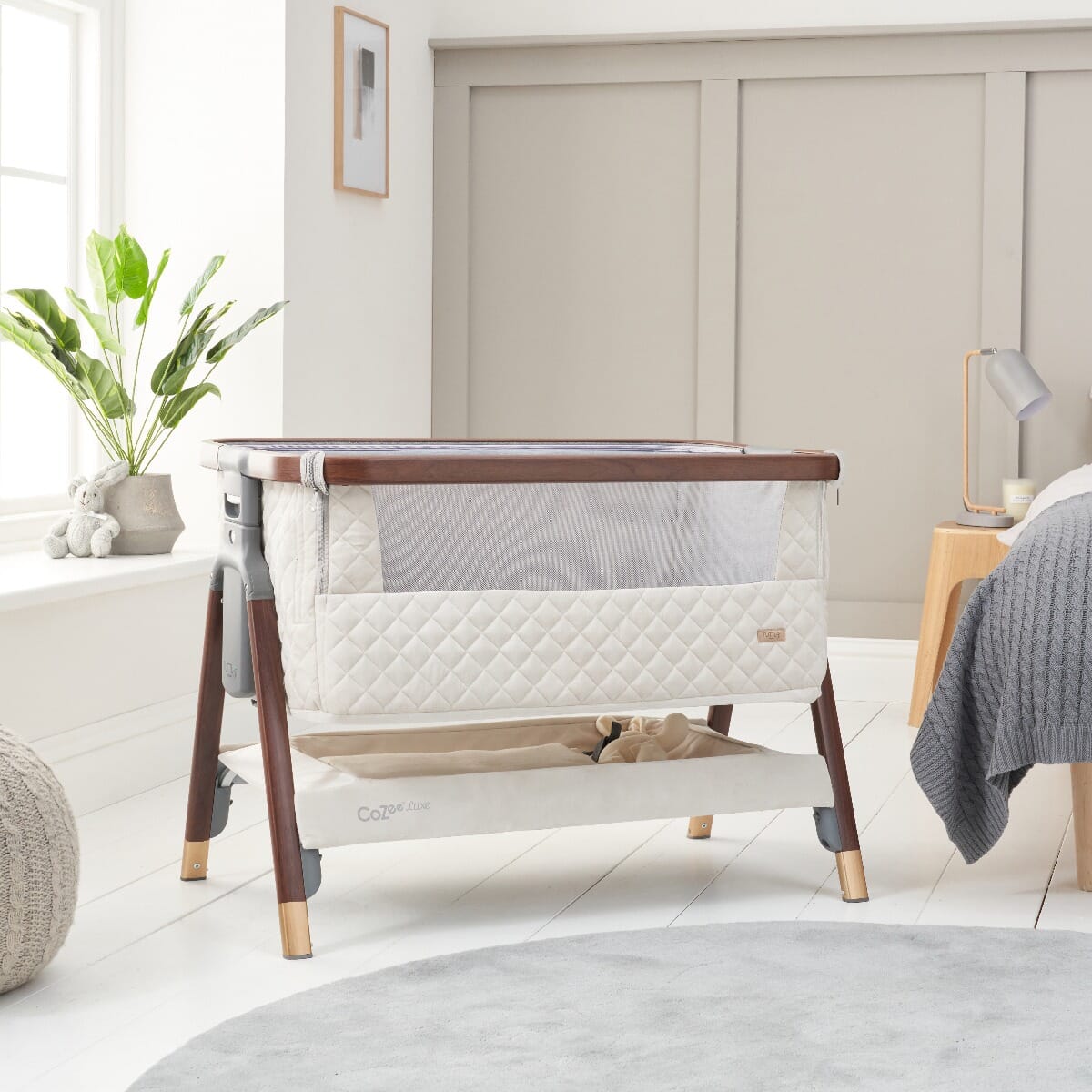 Styled nursery shot of the CoZee Luxe bedside crib in walnut and cream, showcasing breathable mesh sides for visibility and airflow
