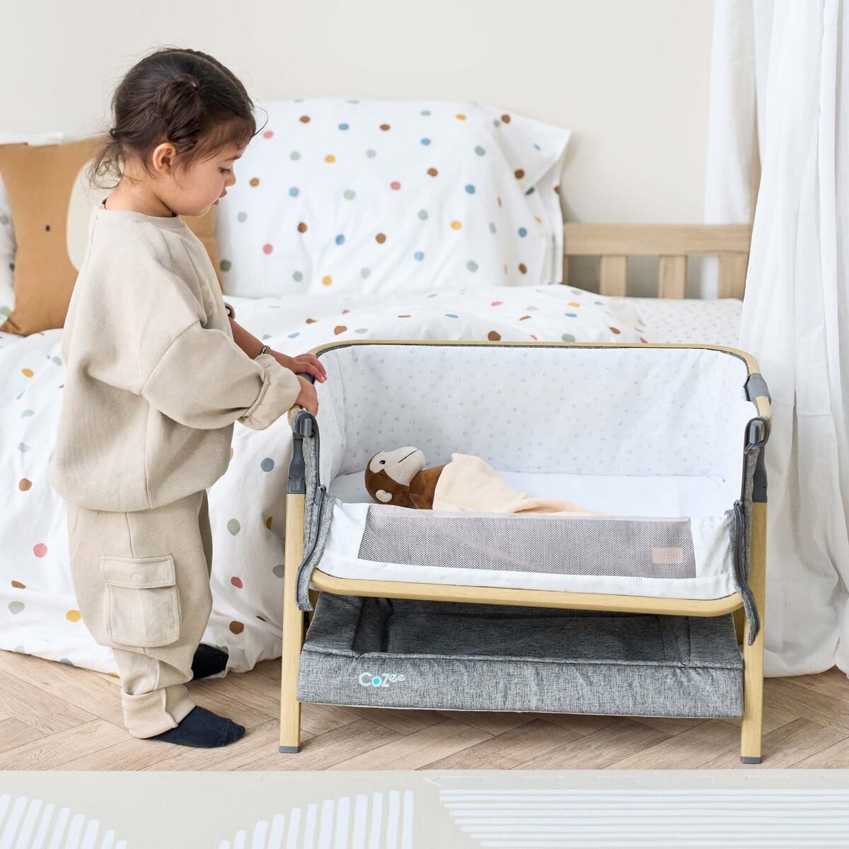 Young child beside the CoZee Mini Me play crib in oak and charcoal, exploring the mesh side which zips down for easy access