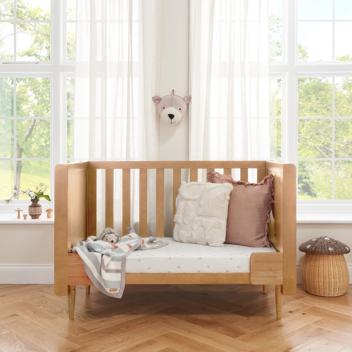Japandi Mini Cot Bed in light oak converted to sofa mode, styled in a bright nursery with soft furnishings. This toddler-friendly setup offers long-term versatility, transitioning from cot to sofa bed for extended use.