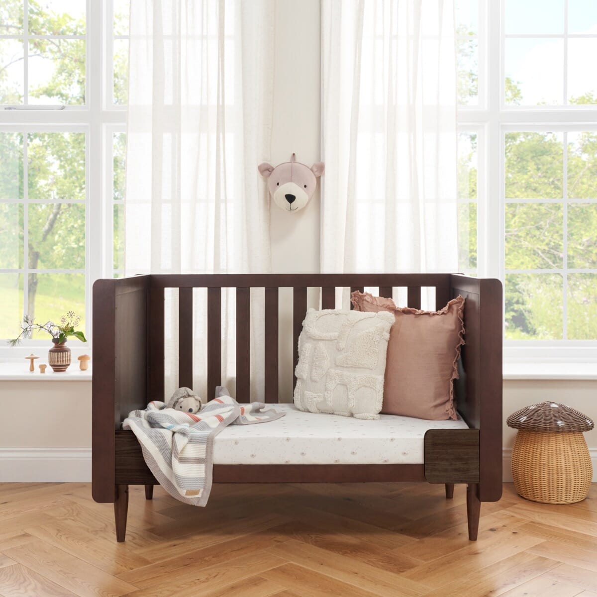 Japandi mini cot bed in warm walnut converted into a toddler sofa, showcasing versatile functionality with supportive sides and plush cushion styling.