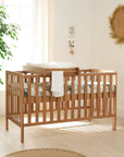 Lifestyle image of the Malmo cot bed in oak with cot top changer, showing practical over-cot setup and natural styling in nursery