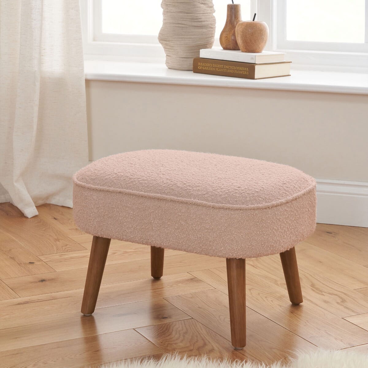 Lifestyle view of Micah Footstool in boucle blush placed by a window, featuring rounded cushioned top and sturdy wooden legs for added comfort and stability.