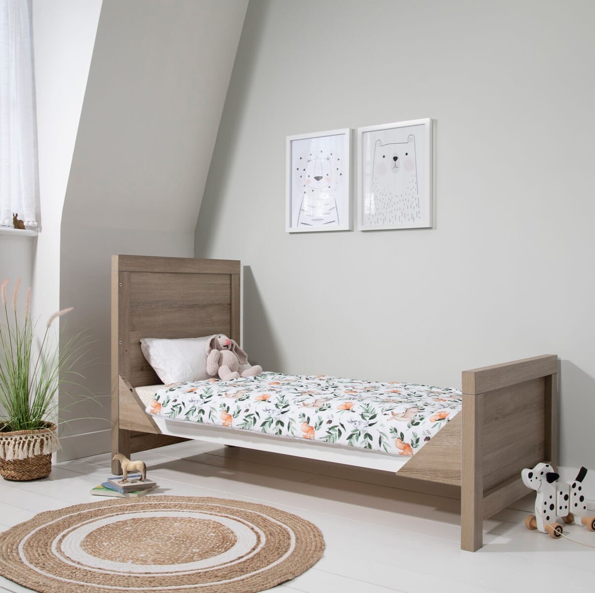 Modena Cot Bed in White and Oak converted into toddler bed mode, styled in a modern nursery with playful bedding and soft toys, offering a practical transition from cot to first bed.