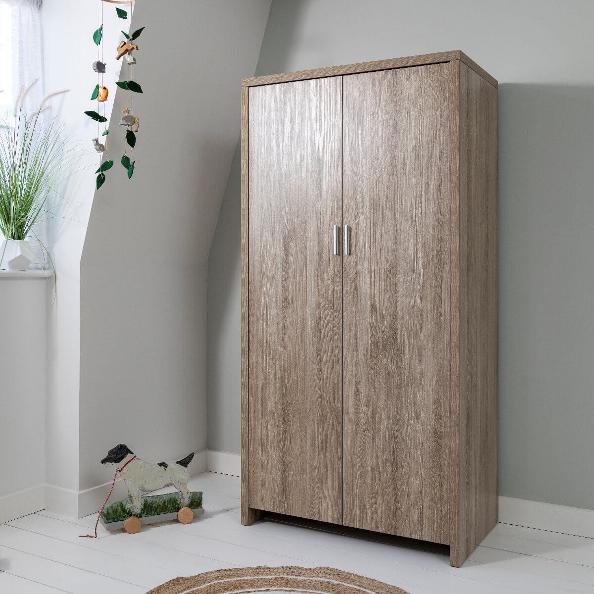Modena wardrobe in oak finish shown in a child's bedroom with doors closed, featuring a warm wood grain texture and compact design for stylish and practical nursery storage.