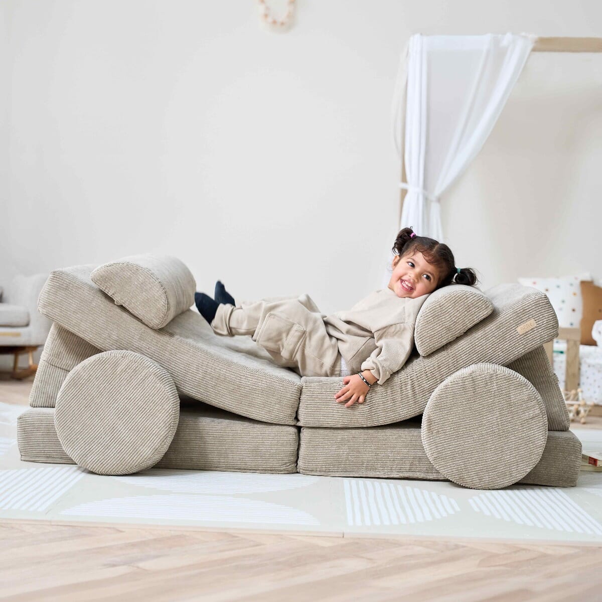 Toddler smiling while lying back on the Playrama Montessori Kids Play Sofa in mushroom finish, arranged into a playful lounger setup
