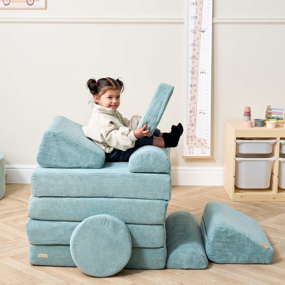 Toddler sitting and smiling while playing with cushions on the Playrama Montessori Kids Play Sofa in ocean stone, styled as a stacked seat