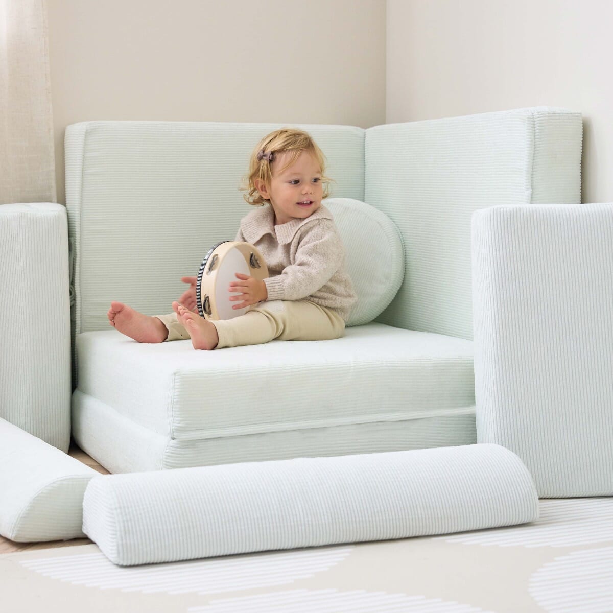 Young girl sits on the Playrama Montessori kids play sofa in sea mist, holding a tambourine, with cushions arranged for cosy play