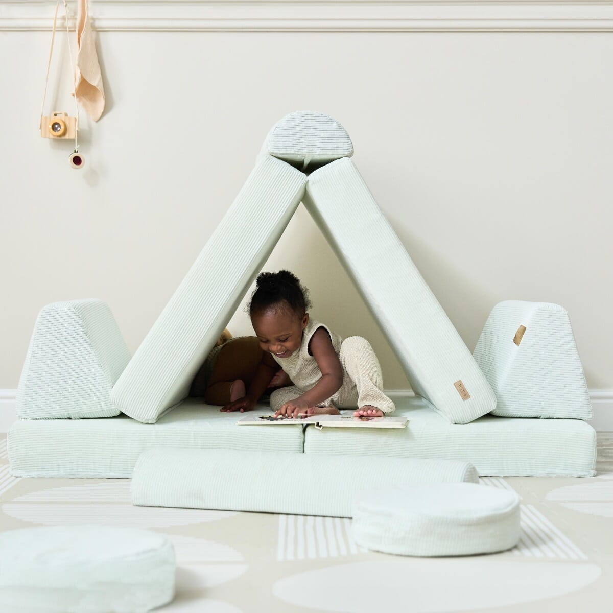 Toddler plays under a tent-shaped build of the Playrama Montessori kids play sofa in sea mist, reading a book inside the cosy setup