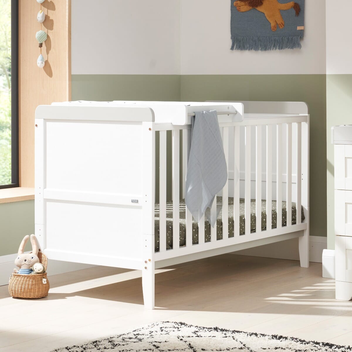 The Rio cot bed in white and dove grey styled in a bright nursery with cot top changer in place, illustrating practical functionality and a neutral palette ideal for any interior.