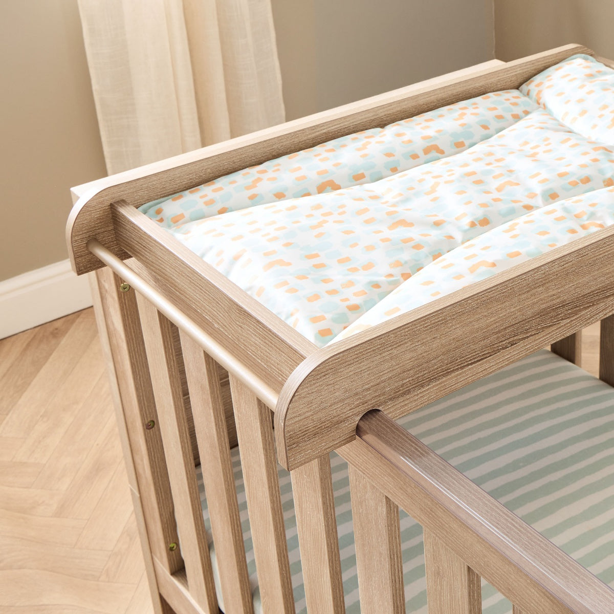 Close-up of the universal cot top changer in oak finish securely fitted to cot bed, featuring raised sides and a leopard print changing mat