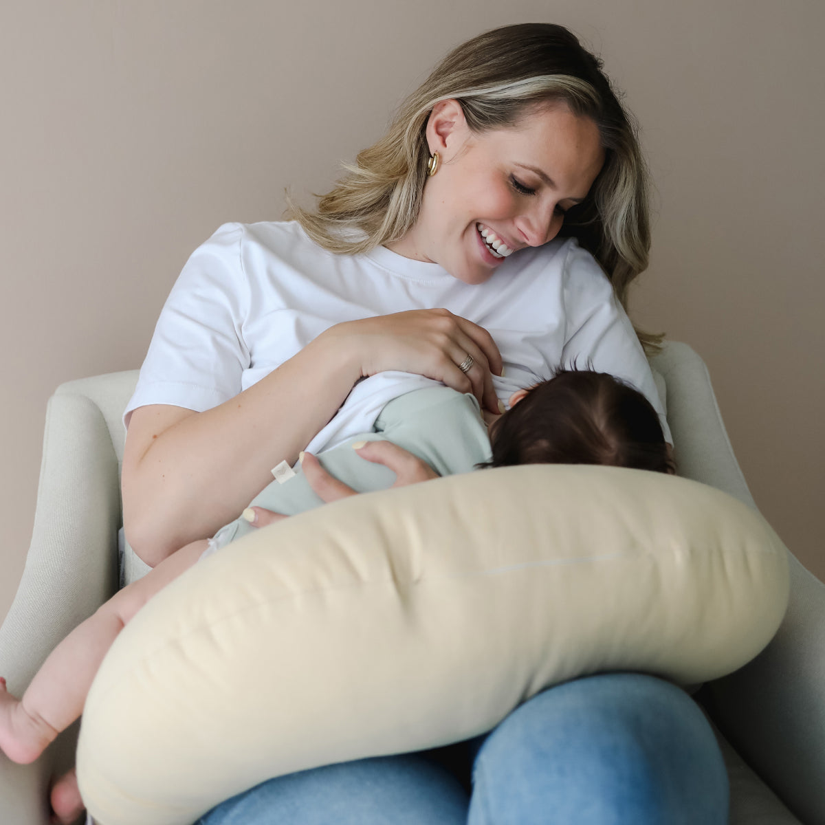 mother smiling whilst holding and breastfeeding baby on a yellow feeding pillow