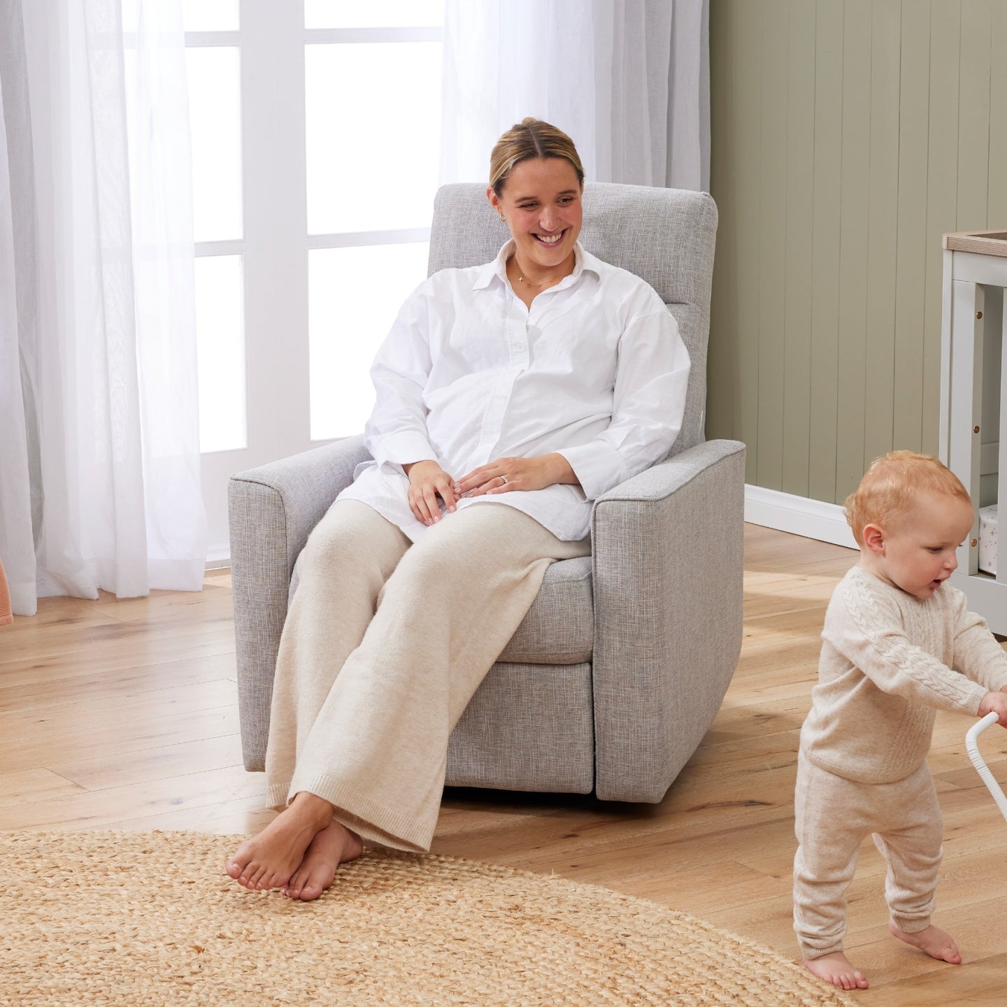 A woman in a white shirt and beige pants is sitting in the grey Paige Recliner Glider nursery chair with a baby standing nearby.