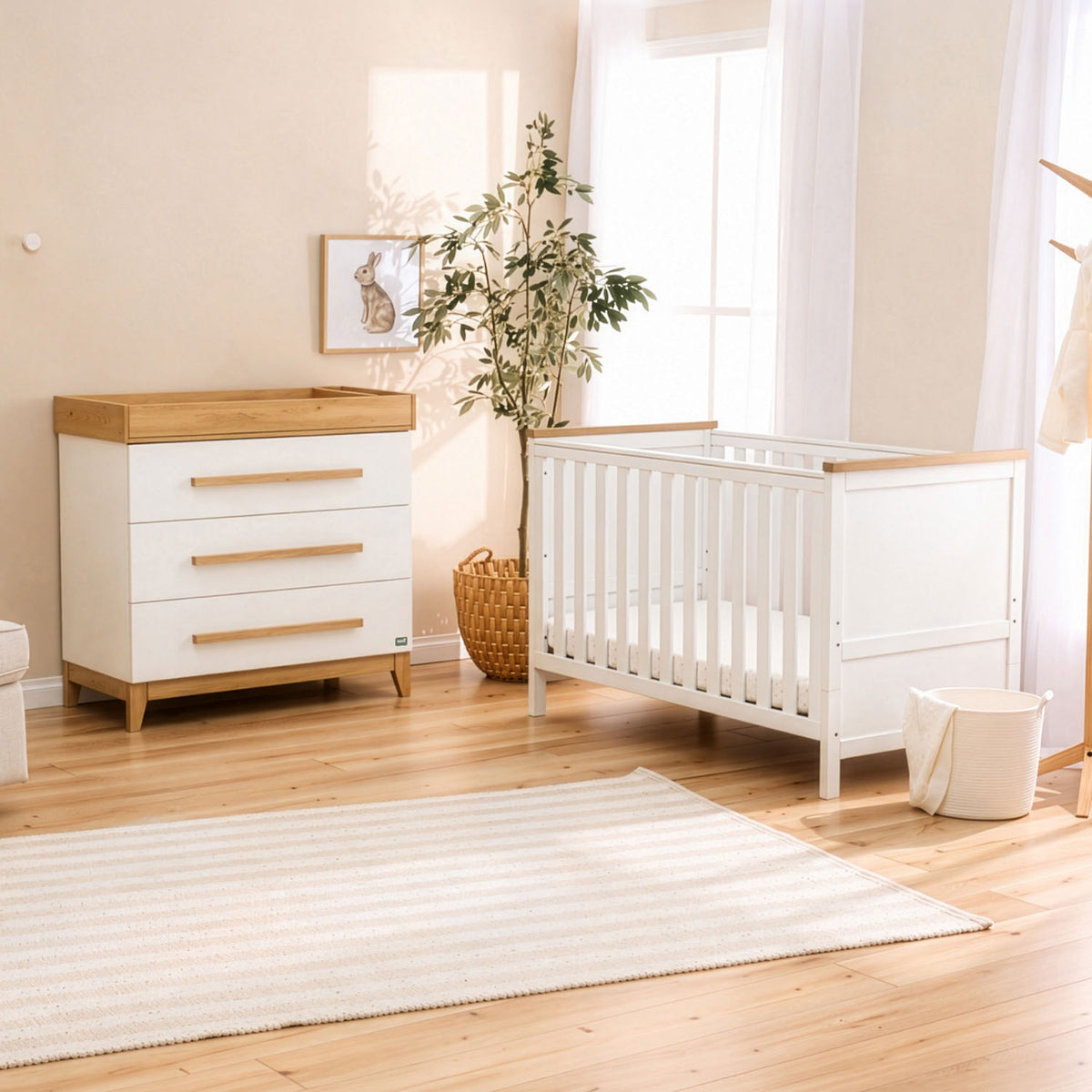 A white wooden baby cot bed with natural golden oak trim stands in a bright nursery next to a matching white chest of drawers with golden oak legs and a top integrated baby-changing unit, a striped rug, and a potted plant.